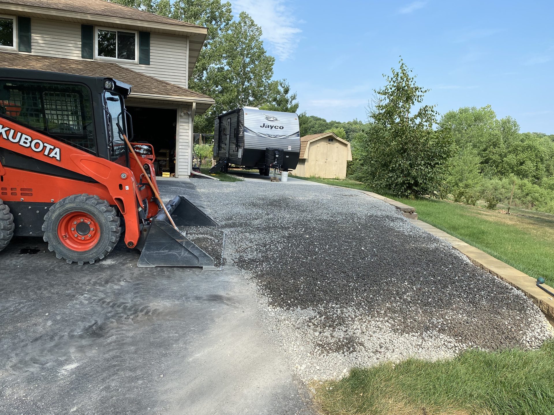 Kubota skid steer spreading gravel on a gravel base under construction, with residential house and Jayco trailer visible in background.