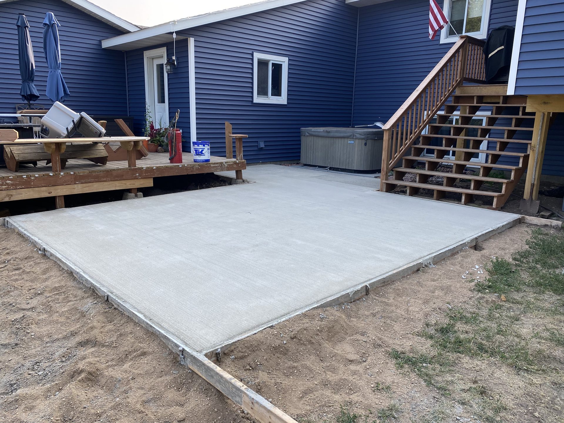 Newly finished concrete patio pad with wooden deck and stairs flanking either side of a blue residential home.