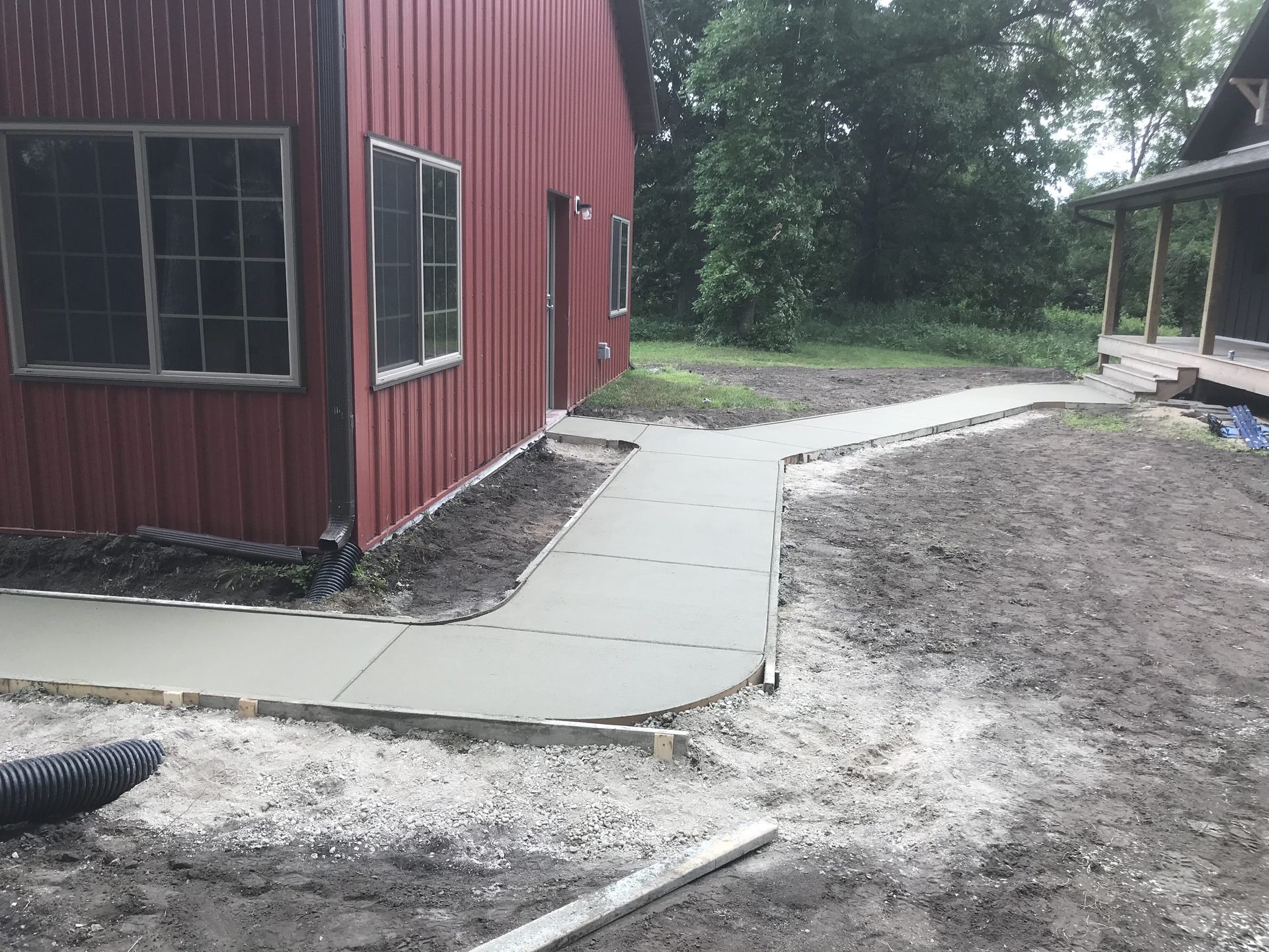 Newly poured concrete walkway and pad beside a red metal building with landscaping soil staging areas visible.