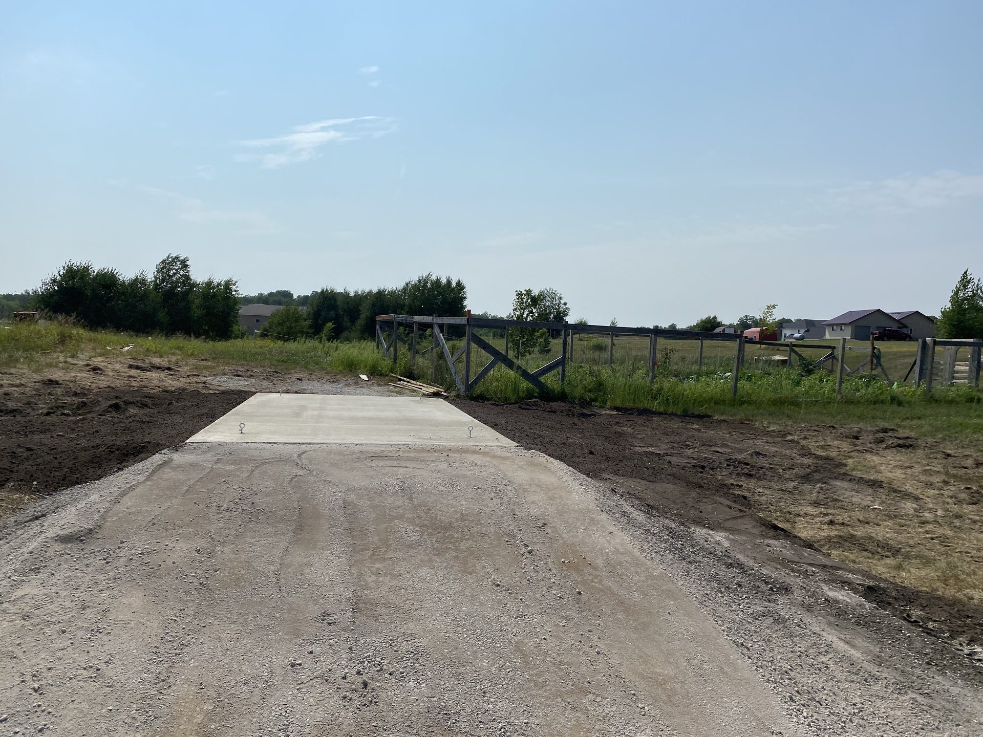 Newly poured concrete pad for driveway entrance on rural property with gravel base and fencing visible.