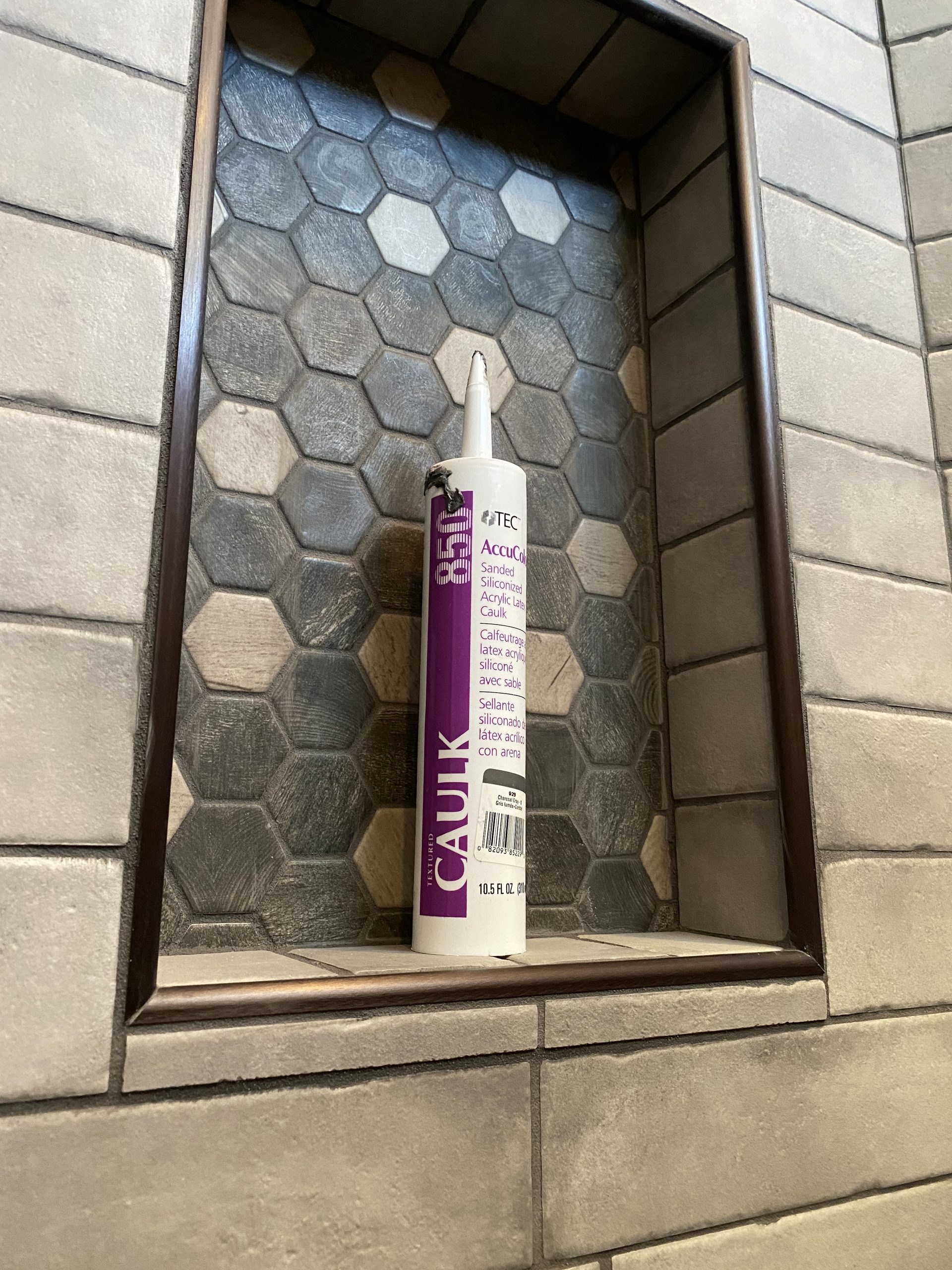 Completed tile shower niche with hexagonal gray tile pattern and white caulk applicator displayed on shelf.